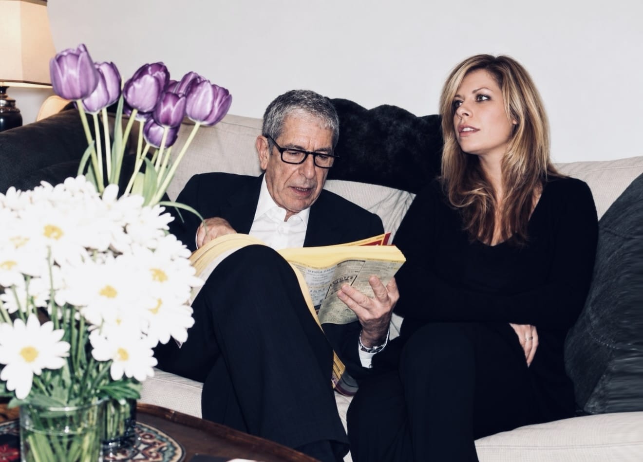 Leonard Cohen and Lonelle Selbo sit on Leonard's couch in his Montreal home while he looks up the number for the chicken place in the phone book.