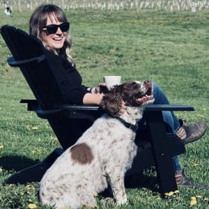 Sara Boyd, Pommelier at Loch Mor Cider sits in a muskoka chair with her dog beside her, overlooking the young orchard.