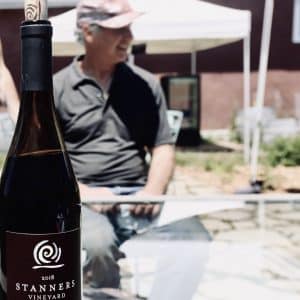 Colin Stanners, Winemaker at Stanners Vineyard, sits at the table outside the winery, a bottle of Pinot Noir in the foreground.