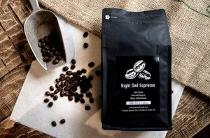 A bag of The Bean Counter Café’s Night Owl Espresso surrounded by roasted coffee beans and a metal scoop.