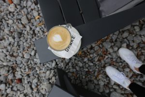 An overhead view of a latte resting on a black chair beside white sneakers on the gravel patio at the Drake Devonshire in Wellington.