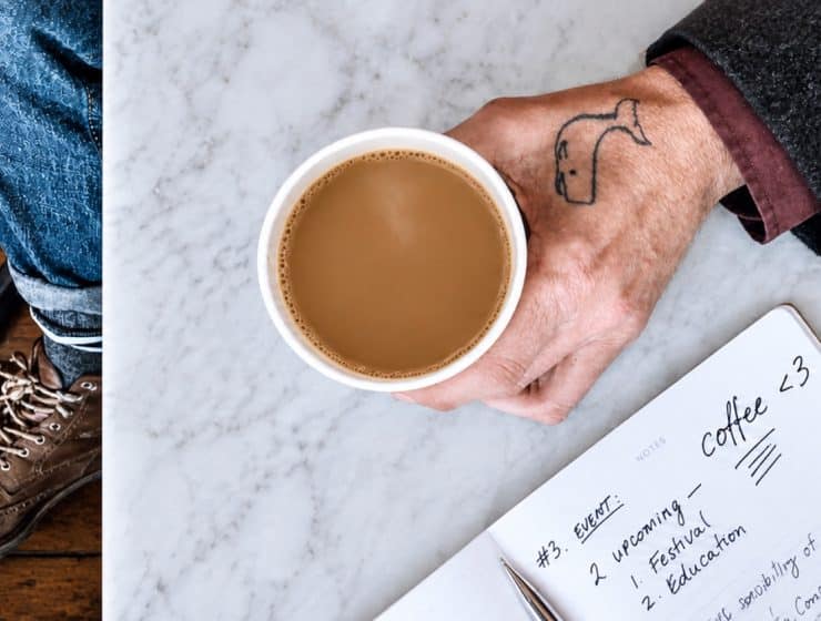 A tattooed hand holds a cup of coffee over a marble café table beside a notebook and worn brown boot.