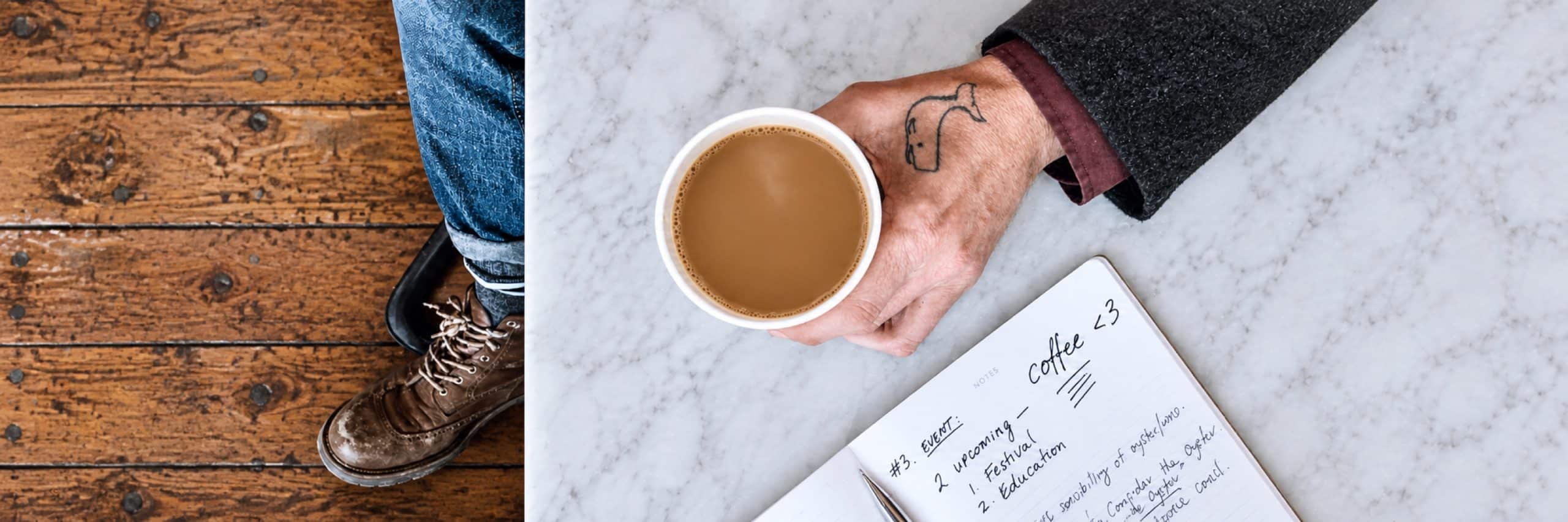 A tattooed hand holds a cup of coffee over a marble café table beside a notebook and worn brown boot.